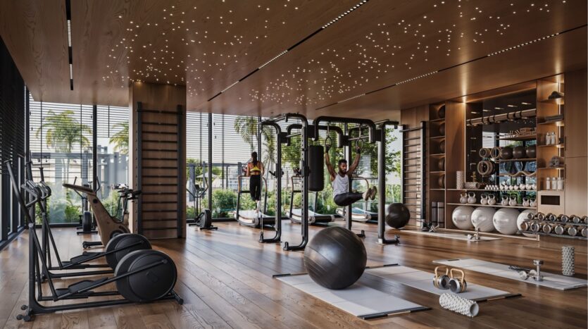Moderno gimnasio en el distrito Damac de Dubai con suelos de madera, grandes ventanales, bicicletas estáticas, pesas libres, colchonetas de yoga y aparatos de fitness. Dos personas se ejercitan mientras la vegetación resplandece en el exterior y las luces del techo crean un efecto de cielo estrellado.