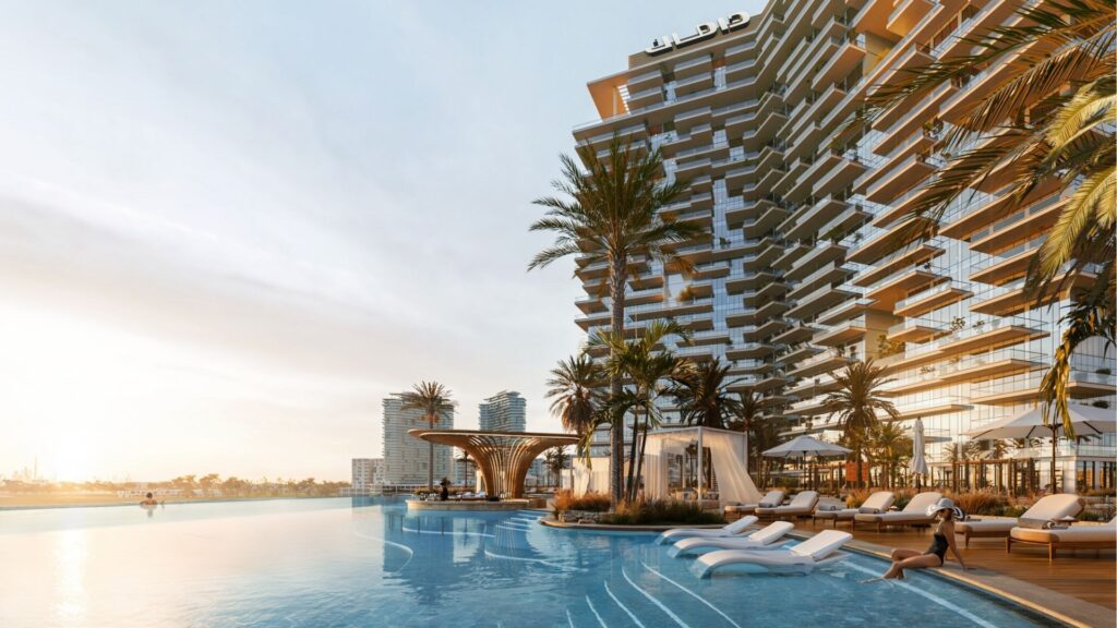 Un lujoso complejo frente al mar en el distrito Damac de Dubai cuenta con modernas terrazas, tumbonas junto a una piscina infinita, palmeras y cabañas al atardecer, con los edificios de la ciudad visibles al fondo.