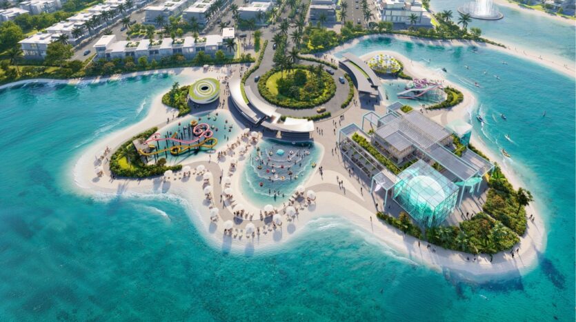 Aerial view of DAMAC Islands 2, a dynamic resort island with pools, waterslides, relaxation areas, umbrellas, modern buildings and palm trees surrounded by clear turquoise water and sandy beaches.