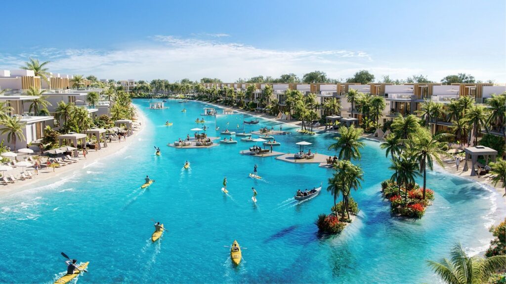 La gran laguna azul claro de Damac Islands 2 está rodeada de modernas villas, palmeras y playas de arena, donde la gente practica kayak, paddleboard y se relaja en plataformas flotantes bajo un brillante cielo azul.