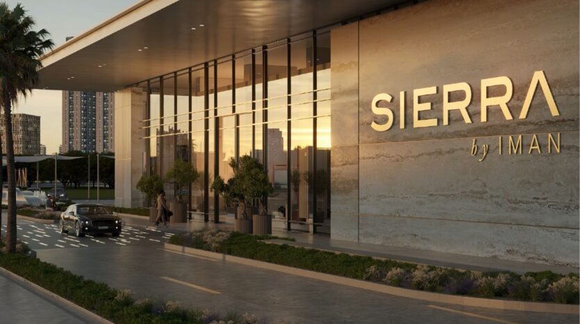Un bâtiment moderne avec une façade en verre et une grande enseigne "sierra by iman" se dresse au milieu d'un paysage verdoyant, d'une voiture à l'entrée et de l'horizon de la ville qui s'illumine au coucher du soleil.