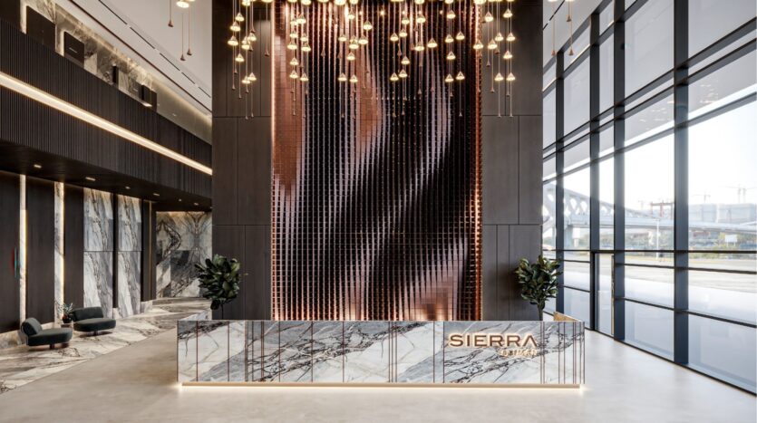 Un hall d'entrée de bureau moderne avec un comptoir de réception en marbre portant l'inscription "SIERRA by Iman", un mur spectaculaire avec des lampes suspendues, de grandes fenêtres et de grandes plantes de chaque côté du comptoir.