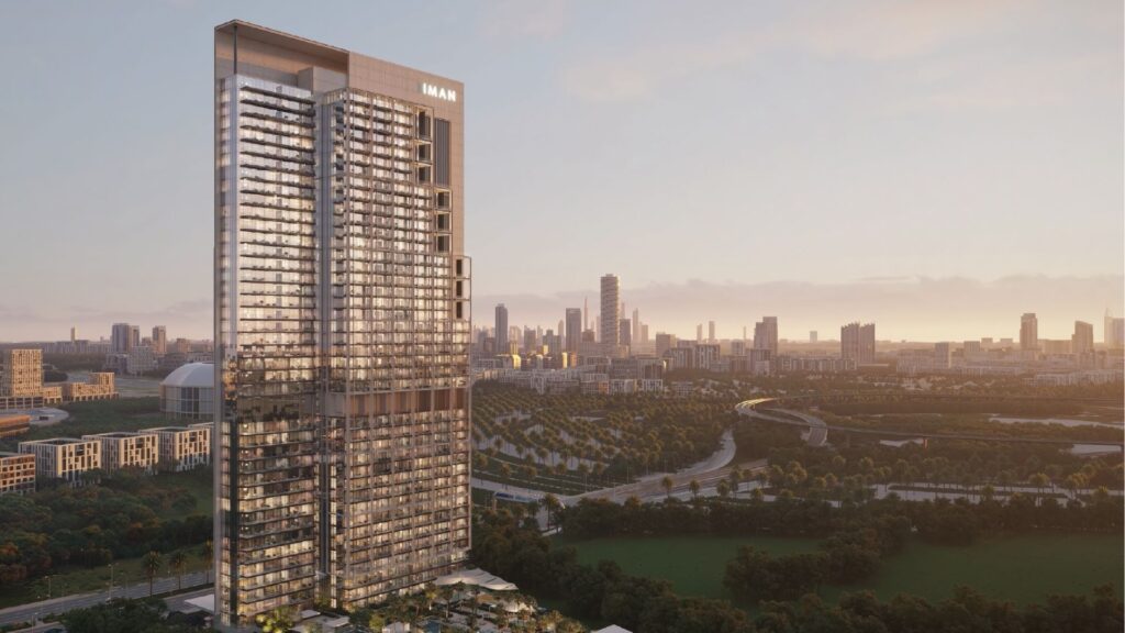 Un grand gratte-ciel moderne portant l'inscription "EMAAR" s'élève au-dessus d'un parc verdoyant, avec le développement Sierra by Iman et l'horizon de la ville en arrière-plan, dans un ciel brumeux au coucher du soleil.