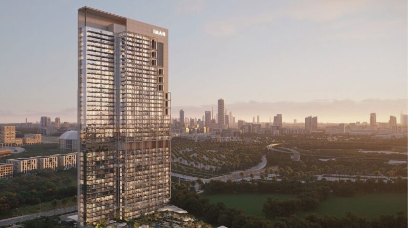 Un grand gratte-ciel moderne portant l'inscription "EMAAR" s'élève au-dessus d'un parc verdoyant, avec le développement Sierra by Iman et l'horizon de la ville en arrière-plan, dans un ciel brumeux au coucher du soleil.