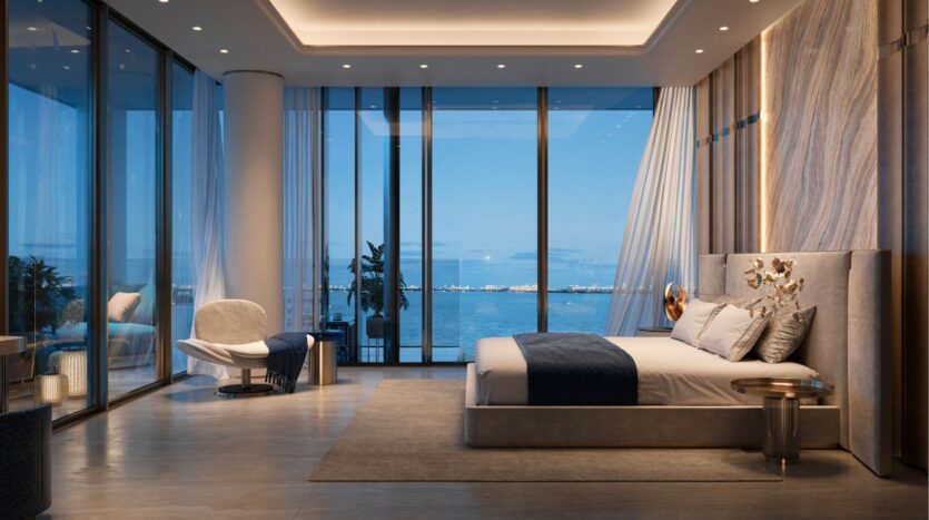 Chambre moderne et luxueuse avec des fenêtres allant du sol au plafond et donnant sur la mer calme et bleue des îles Silena Dubaï. La chambre comprend un grand lit avec des draps bleus et blancs, un fauteuil en peluche, un éclairage élégant et un décor minimaliste.