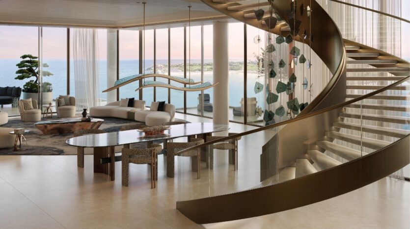 Un espace de vie moderne et spacieux avec des fenêtres allant du sol au plafond et offrant une vue sur l'océan et les îles Silena Dubai, un élégant escalier incurvé, un mobilier contemporain et une grande table à manger, le tout baigné de lumière naturelle.