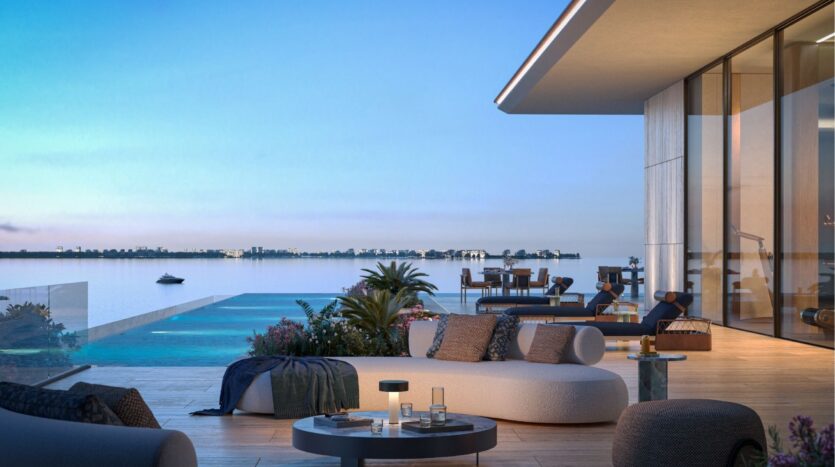 Une terrasse extérieure moderne avec des sièges élégants, une piscine surplombant une étendue d'eau calme et un bâtiment aux murs de verre au coucher du soleil - l'élégance des îles Silena Dubaï. Un bateau flotte au loin sous un ciel bleu clair.