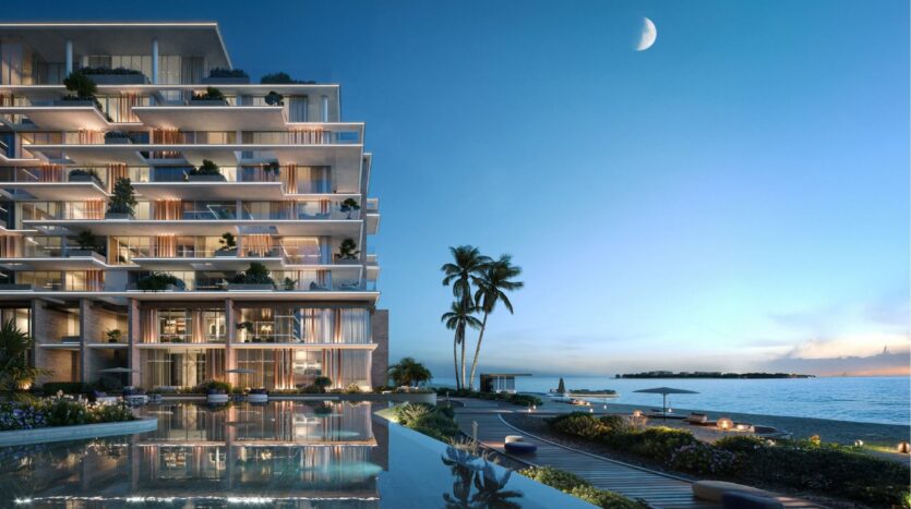 Un bâtiment moderne en bord de mer à Silena Dubai Islands dispose de balcons en verre donnant sur une piscine calme, des palmiers et une plage de sable au crépuscule, sous un croissant de lune. Le ciel passe du bleu profond au pâle le long de l'horizon.