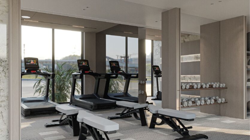 Une salle de sport moderne avec trois tapis de course face à de grandes fenêtres, plusieurs bancs d'entraînement blancs et noirs et des étagères remplies d'haltères argentés. La lumière naturelle pénètre dans l'espace par les fenêtres.