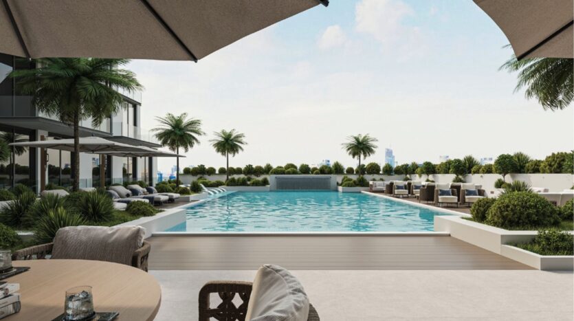 Une piscine extérieure moderne avec des chaises longues, des parasols et des palmiers. Au premier plan, des tables et des chaises, des buissons bien entretenus et la silhouette d'une ville visible au loin sous un ciel dégagé.