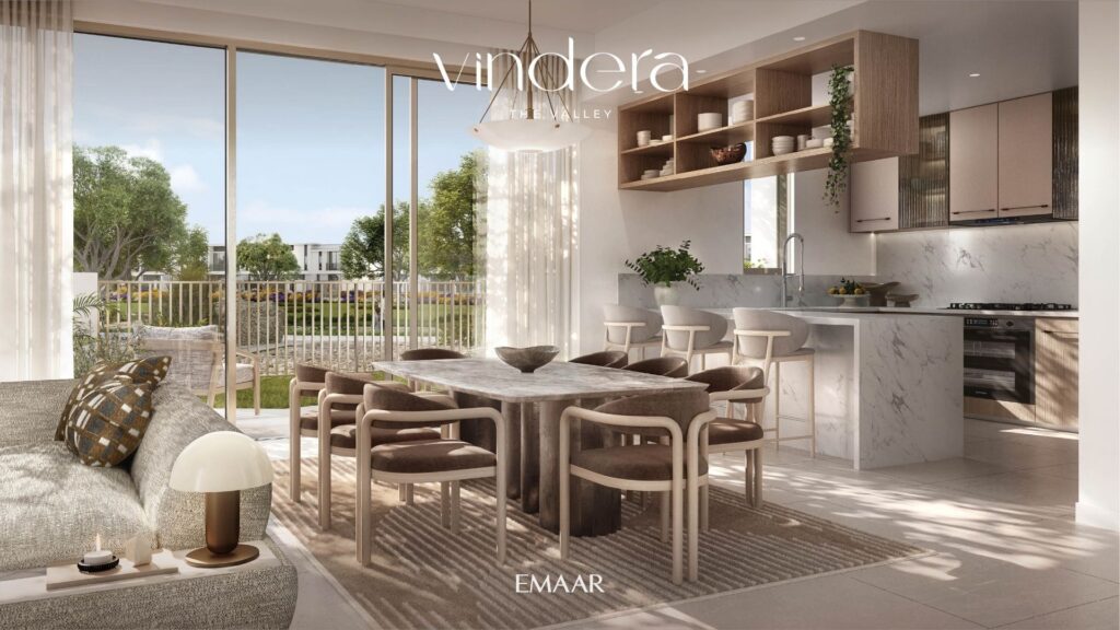 A sunny, modern living and dining room, with marble kitchen, wooden dining table, beige chairs, large windows and a view of lush outdoor space. The words "Vandera, The Valley" and "EMAAR" are displayed on the image.