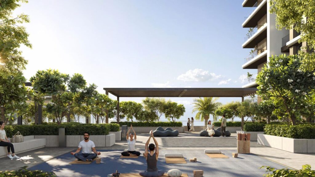 Le persone praticano yoga all'aperto su tappetini in un moderno cortile paesaggistico del Lyvia by Palace Residence nel porto di Dubai Creek, circondato dal verde e da alti edifici, mentre altri si rilassano o passeggiano sullo sfondo sotto un limpido cielo blu.