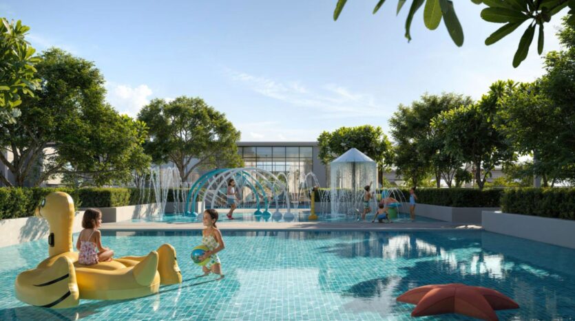 Bambini giocano in una piscina con una papera gonfiabile gialla al Lyvia by Palace Residence nel Dubai Creek Harbour, circondato da alberi e vegetazione. Altri bambini si godono le fontane e le strutture di gioco sullo sfondo in una giornata di sole.
