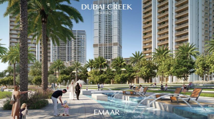 Grattacieli moderni e palme circondano un parco paesaggistico con una fontana, lettini e persone che si rilassano nel porto di Dubai Creek, vicino al Lyvia by Palace Residence. Un uomo aiuta un bambino in riva al mare mentre altri si godono la giornata di sole.