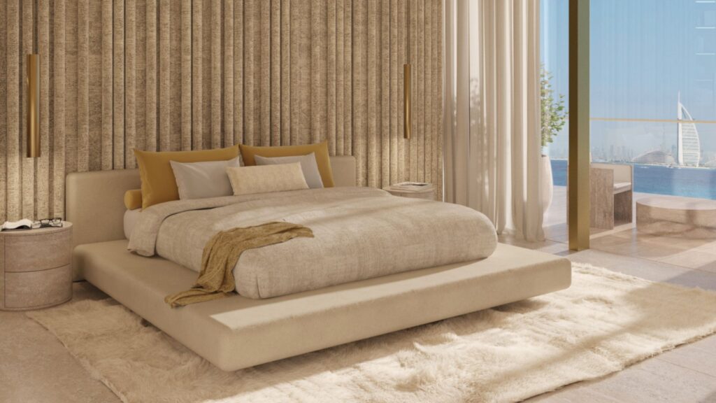 В современной спальне Maison Margiela Residences бежевая кровать-платформа, нейтральное многослойное постельное белье, золотые акцентные подушки и фактурная стена. Окна от пола до потолка выходят на балкон с видом на океан и Бурдж Аль Араб вдали.