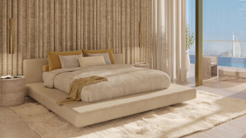 В современной спальне Maison Margiela Residences бежевая кровать-платформа, нейтральное многослойное постельное белье, золотые акцентные подушки и фактурная стена. Окна от пола до потолка выходят на балкон с видом на океан и Бурдж Аль Араб вдали.