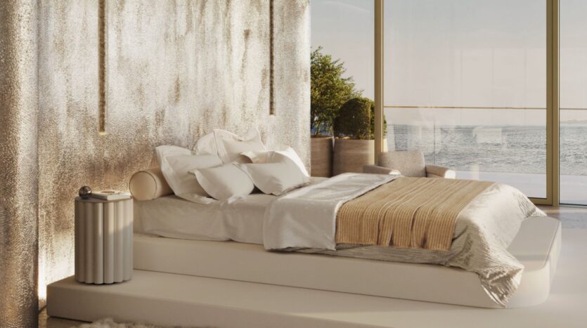 В современной спальне Maison Margiela Residences низкая белая кровать, белое и бежевое постельное белье, фактурная металлическая стена, окна от пола до потолка с видом на воду и зелень, а также серое кресло в углу.