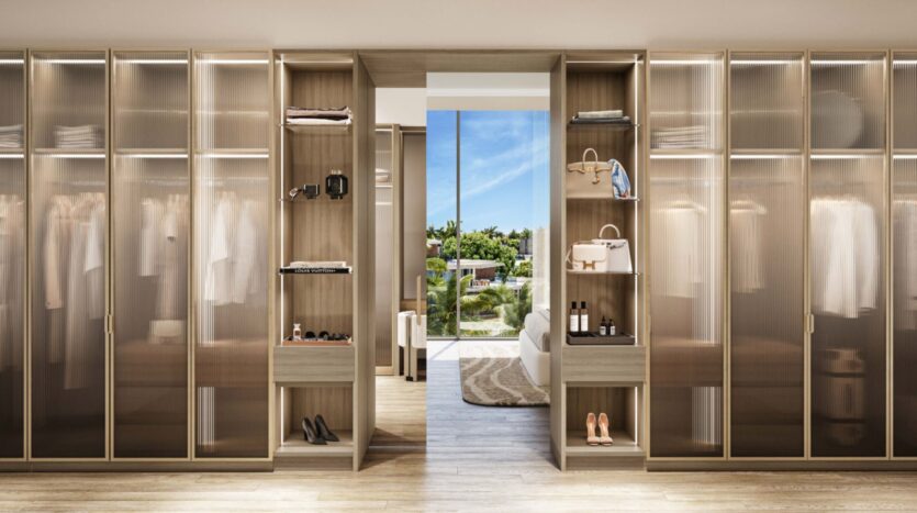 Современная гардеробная Palm Central со стеклянными шкафами, в которых аккуратно хранятся одежда, обувь и аксессуары. Открытая дверь открывает светлую спальню и вид на зелень и голубое небо за окном.