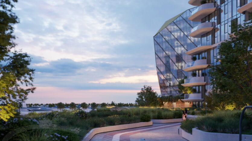 Un bâtiment moderne en verre avec des balcons incurvés surplombe le Portside Square Mina Rashid, avec une allée paysagère bordée de verdure et d'arbres. Dans un ciel de coucher de soleil, deux personnes se promènent sur le sentier panoramique.