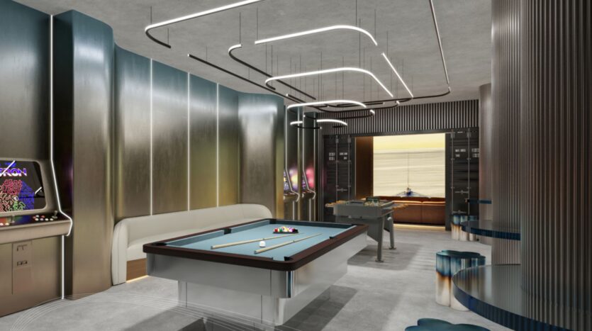 La salle de jeux moderne du Portside Square Mina Rashid comprend un billard, des machines d'arcade, un baby-foot, des murs métalliques élégants, des plafonniers uniques et des meubles contemporains, dont un canapé blanc et des tabourets aux tons bleus.