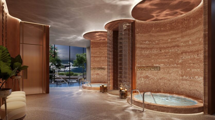 Salle de spa luxueuse dotée d'un éclairage chaleureux, de murs texturés, de deux jacuzzis ronds, d'un jeu d'eau décoratif, de bougies, de chaises longues, de plantes luxuriantes et d'une grande fenêtre donnant sur la piscine et la verdure au Portside Square Mina Rashid.