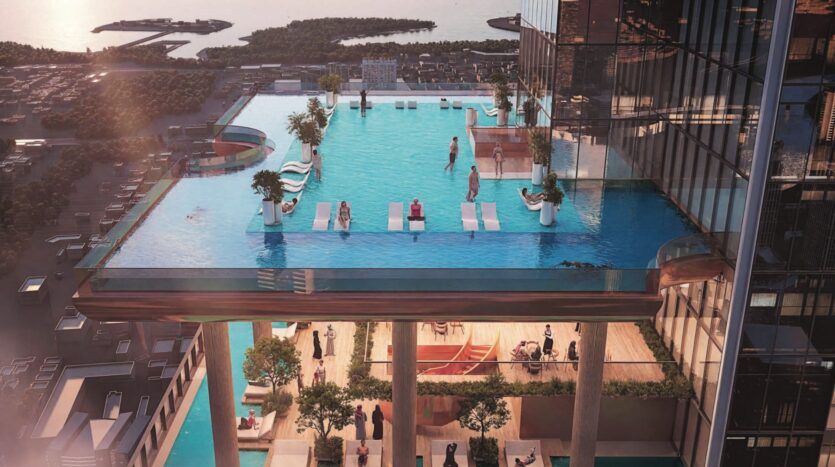 A rooftop infinity pool, with sun loungers and people relaxing at the SkyParks, overlooking a cityscape, water and distant islands. The pool's glass walls crown a modern high-rise building with a terrace below.