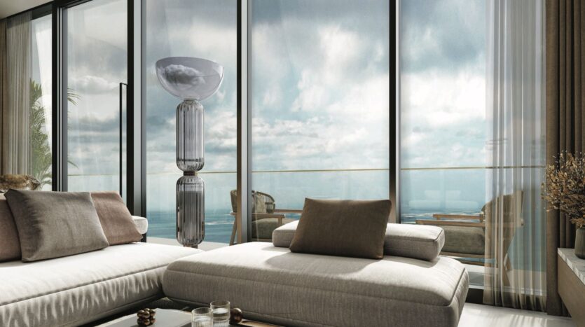 Modern living room with beige sofas and neutral cushions, large floor-to-ceiling windows, a tall lamp and a view of an outdoor SkyParks balcony overlooking the ocean under cloudy skies.