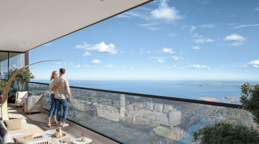 A couple stands on a modern SkyParks balcony overlooking a cityscape and a vast ocean under a clear blue sky, surrounded by elegant outdoor furniture and green plants.