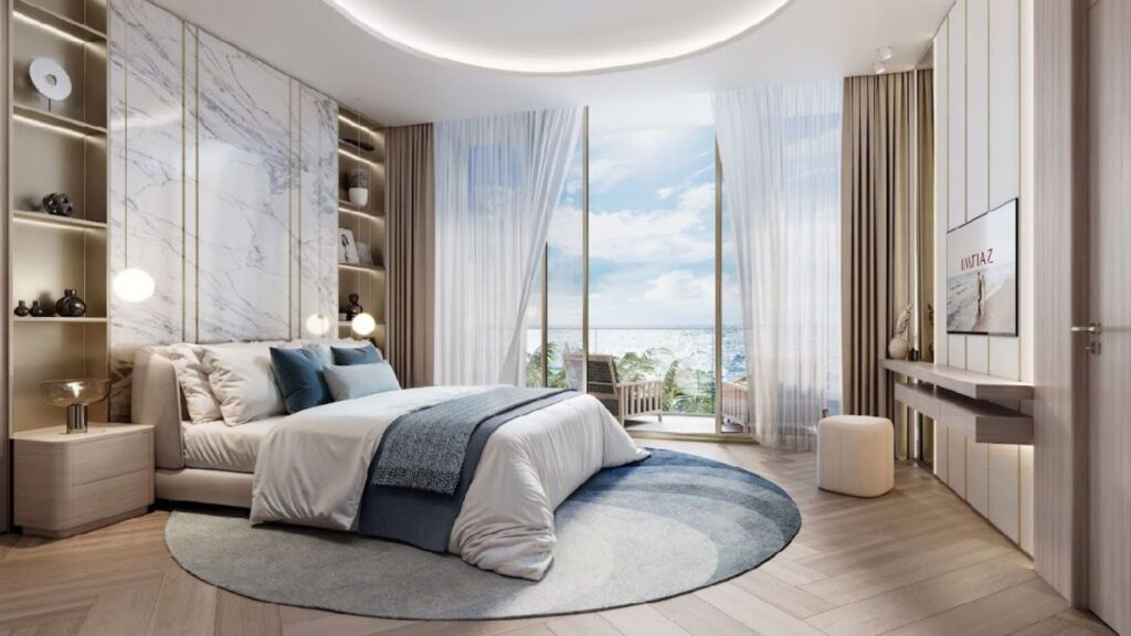 Una moderna camera del Sunset Bay Grand Residence by Imtiaz a Dubai Islands è dotata di letto king-size, biancheria da letto blu e bianca, moquette circolare, finestre a tutta altezza, tende trasparenti, vista sull'oceano e lussuosi tocchi di marmo e legno.