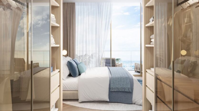 Camera da letto moderna con toni neutri, un letto ben fatto con lenzuola bianche e blu, scaffali dell'armadio aperti e grandi finestre con vista sull'oceano al Sunset Bay Grand Residence by Imtiaz a Dubai Islands. Tende trasparenti incorniciano la vista serena.