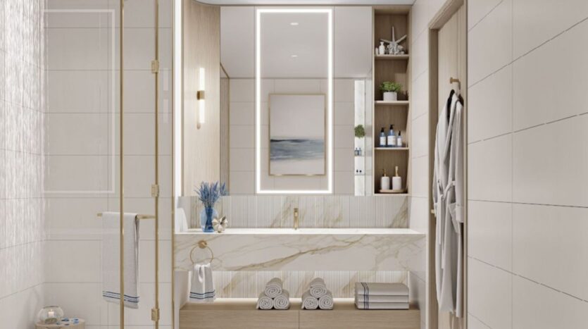 Bagno moderno al Sunset Bay Grand Residence by Imtiaz a Dubai Islands con grande specchio illuminato, piano di lavoro in marmo, scaffali aperti con asciugamani e articoli da toilette, doccia in vetro e piastrelle bianche. Un accappatoio è appeso alla parete, riflesso da opere d'arte incorniciate.