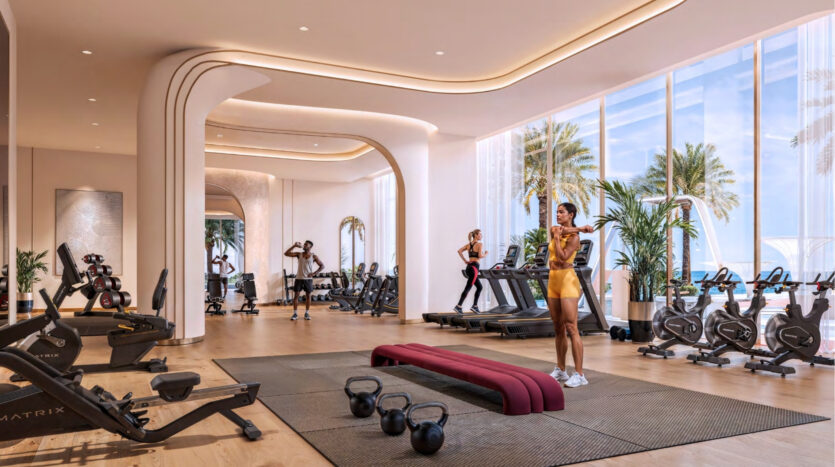 A modern gym at Breez by Danube Maritime City with large windows overlooking palm trees and the ocean, with exercise bikes, treadmills, weights, and several people working out in a bright, spacious environment with natural light.