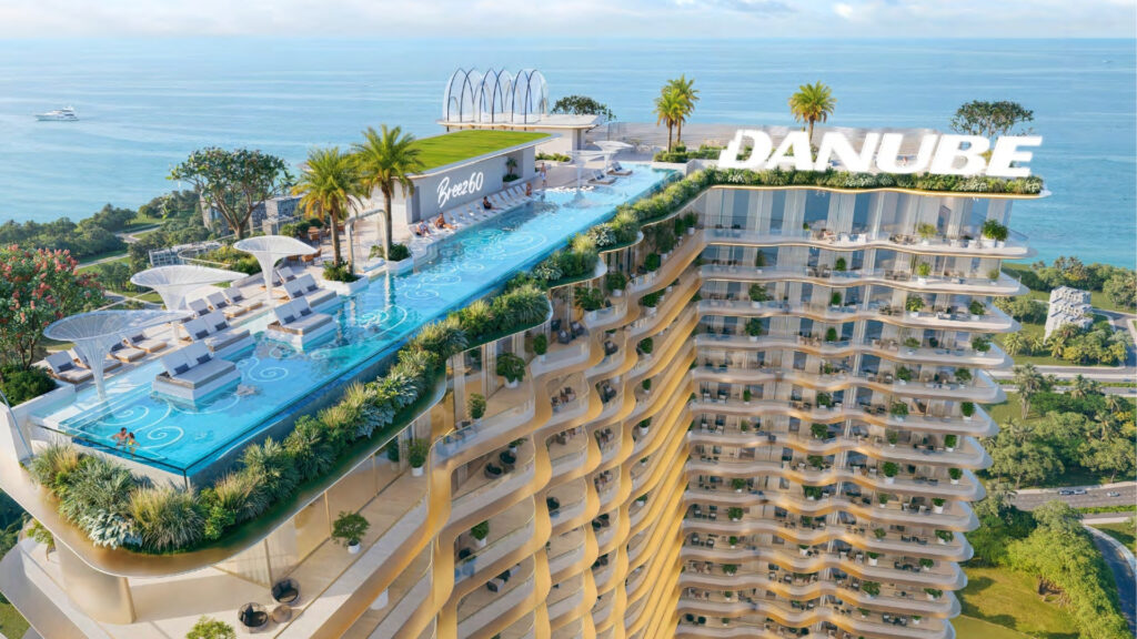 Breez by Danube Maritime City is a luxury tower featuring a rooftop infinity pool, sun loungers, palm trees and lush vegetation overlooking the ocean. The word "DANUBE" appears in large letters at the top of the building.