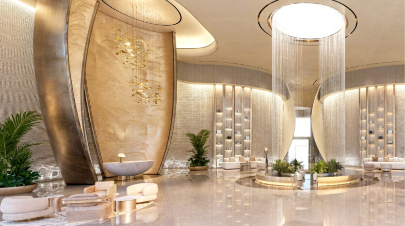 Luxurious lobby with high ceilings, elegant gold and beige décor, curved wall elements, modern seating and lush green plants, reflecting the sophisticated ambience of Breez by Danube Maritime City. A large circular chandelier serves as the centerpiece.