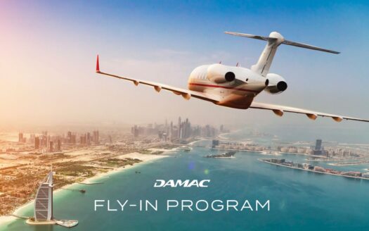 Un jet privé survole la côte de Dubaï avec les gratte-ciel de la ville et le Burj Al Arab en contrebas. Le ciel est dégagé et le texte indique "DAMAC Fly-In Program".