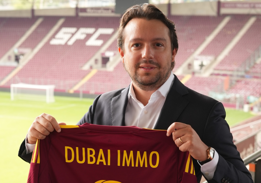 Ein Mann im Anzug hält ein weinrotes Fußballtrikot mit dem Aufdruck "DUBAI IMMO" in die Höhe, in einem Stadion mit leeren weinroten Sitzen und einem Fußballtor, wodurch die Verbindung zur Immobilienbranche in Dubai betont wird.