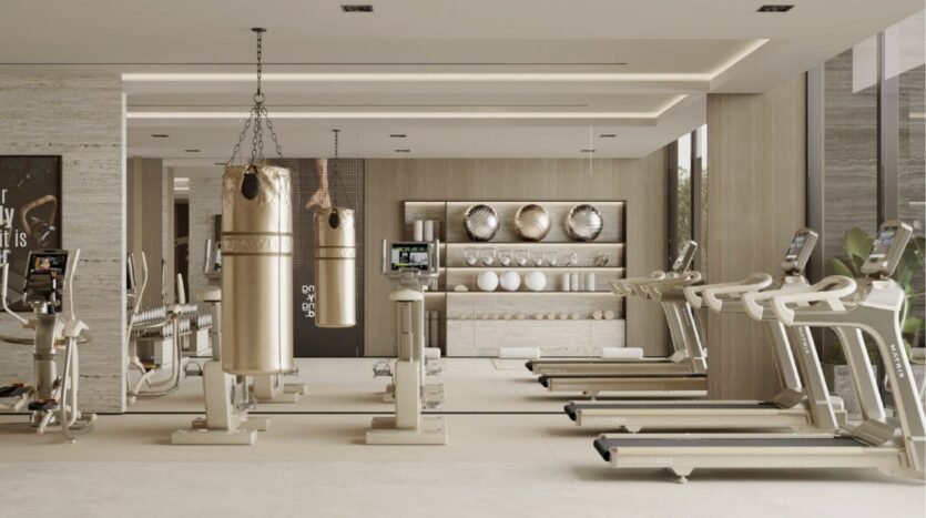 Salle de sport moderne aux tons beiges, équipée de tapis de course, de vélos d'appartement, de sacs de frappe, d'étagères à poids et d'appareils fixés au mur. Inspiré par le Franck Muller Yachting à Dubai Maritime City, l'espace est bien éclairé par de grandes fenêtres et un décor minimaliste.