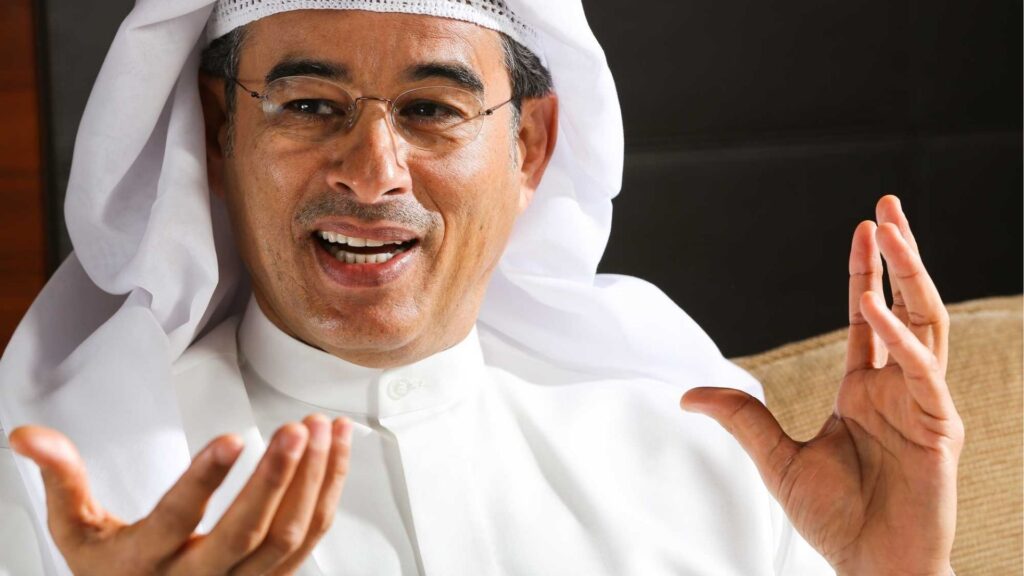 Ein Mann mit einer Brille, einem traditionellen weißen Hut und einem Kopftuch aus dem Nahen Osten sitzt auf einem beigen Stuhl. Er lächelt und macht Gesten mit beiden Händen, während er über die Biografie von Mohamed Ali Alabbar spricht.