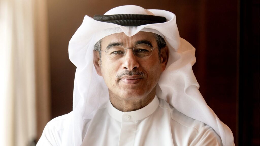 Ein Mann mittleren Alters mit Brille, einer weißen Kandura und einem weißen traditionellen Kopftuch steht darin und lächelt sanft in die Kamera - ein Bild, das an die Biografie von Mohamed Ali Alabbar erinnert. Ein warmes, natürliches Licht und ein verschwommener brauner Hintergrund sind zu sehen.