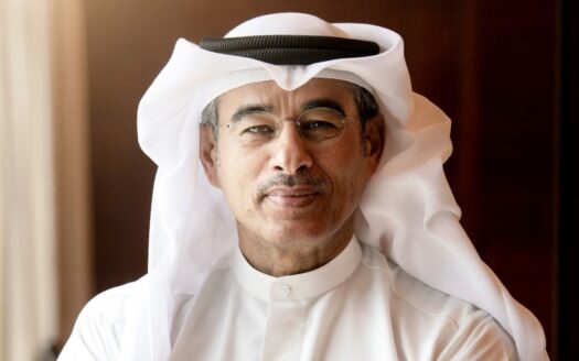 Un homme d'âge moyen portant des lunettes, une kandoura blanche et un foulard traditionnel blanc se tient à l'intérieur, souriant doucement à l'appareil photo - une image qui rappelle la biographie de Mohamed Ali Alabbar. Une lumière chaude et naturelle et un arrière-plan brun flou sont visibles.