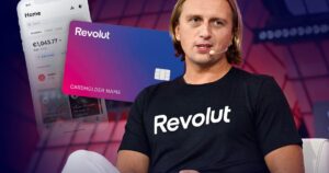Una persona che indossa una maglietta nera "Revolut" parla sul palco attraverso un auricolare. Alle sue spalle scorrono le immagini di una carta Revolut e di uno smartphone che visualizza l'applicazione, mentre apprendiamo che il fondatore della banca Revolut si sta trasferendo negli Emirati.