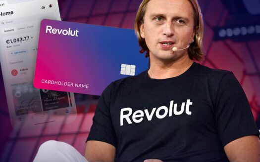 Una persona con una camiseta negra de "Revolut" habla en el escenario a través de unos auriculares. Detrás aparecen imágenes de una tarjeta Revolut y un smartphone que muestra la aplicación, mientras nos enteramos de que el fundador del banco Revolut se traslada a los Emiratos.