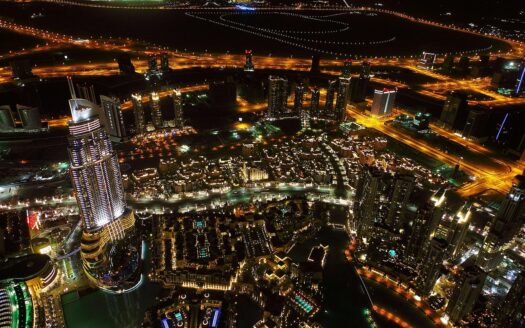 Vue aérienne nocturne d'un paysage urbain très éclairé avec de hauts gratte-ciel, des routes sinueuses et des bâtiments illuminés - parfait pour ceux qui recherchent une location à Dubaï avec une villa luxueuse ou un appartement moderne au milieu des lampadaires lumineux et des espaces ouverts sombres.