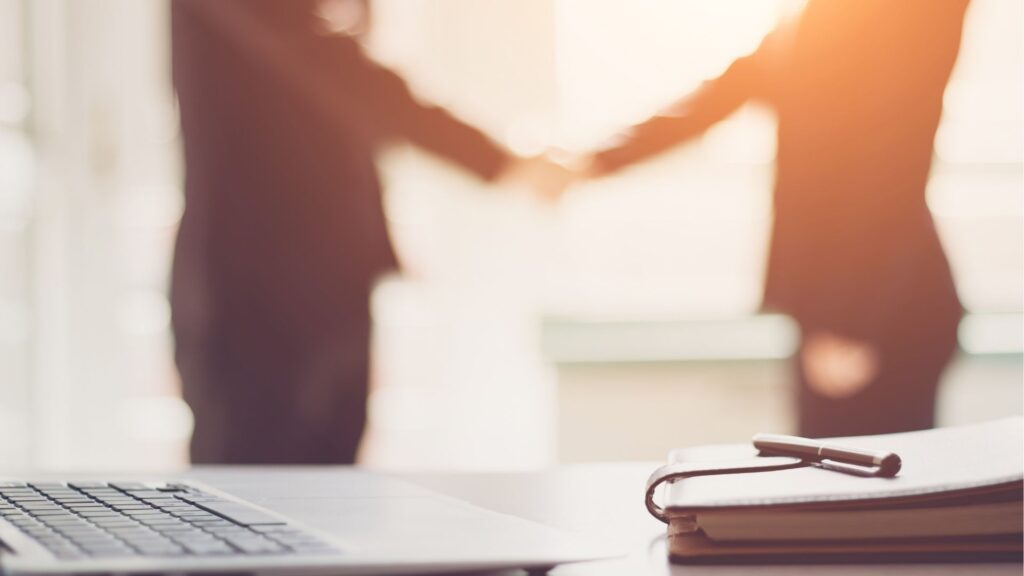 Au premier plan, un bureau avec un ordinateur portable et un bloc-notes ; à l'arrière-plan flou, deux personnes en tenue d'affaires se serrent la main - peut-être pour sceller un contrat d'immobilier à Dubaï - dans un bureau éclairé par le soleil.