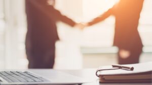 In primo piano, una scrivania con un computer portatile e un blocco note; sullo sfondo sfocato, due persone in abiti da lavoro si stringono la mano - forse per concludere un affare immobiliare a Dubai - in un ufficio illuminato dal sole.