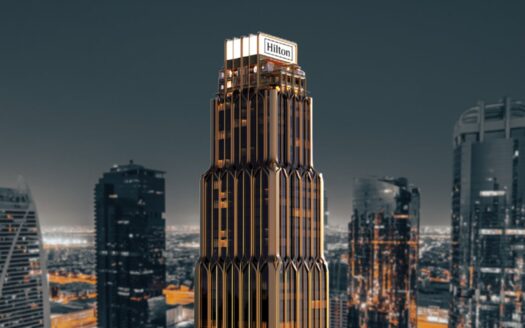 Un gran rascacielos moderno coronado por el letrero "Hilton", las residencias Hilton destacan sobre un paisaje urbano de rascacielos al atardecer, con llamativos acentos dorados y oscuros.