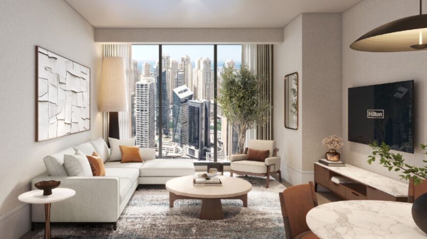 Modern living room from Hilton Residences, featuring a beige sofa, armchair, coffee table and TV. The large window allows you to admire the city skyline with its tall buildings, complemented by an indoor tree and neutral-toned decor.