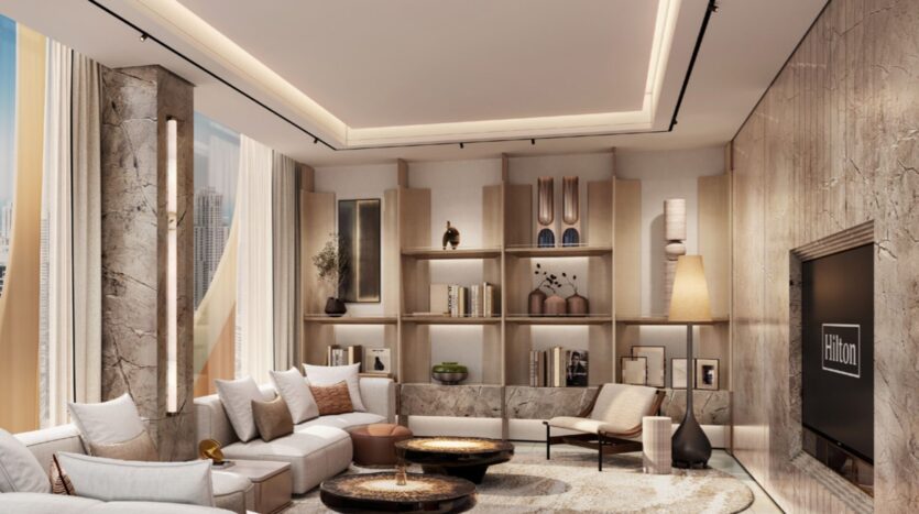 The modern living room at Hilton Residences features a large beige sectional sofa, round coffee tables and built-in shelves for storing books and decorative items. Marble accents, tall windows and neutral tones create a sophisticated, comfortable atmosphere.