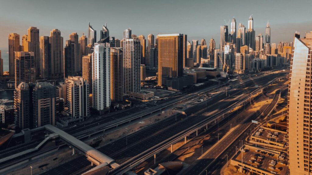 A cityscape featuring numerous buildings and skyscrapers, including elegant Hilton residences, with a multi-lane highway running through the center, bathed in warm sunlight under clear skies.