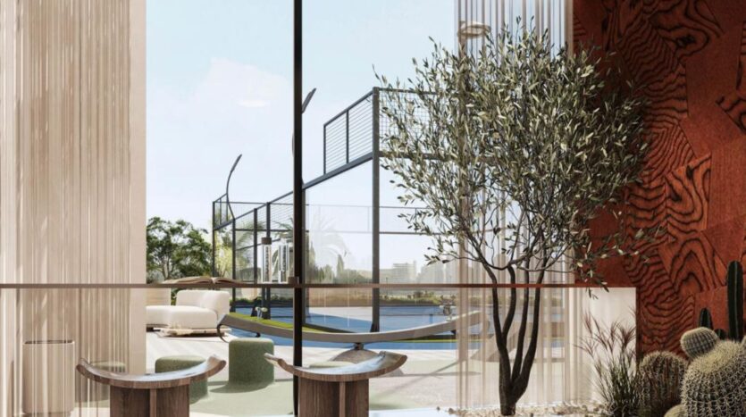 Modern interior space in Rabdan Gardens with large windows overlooking an outdoor tennis or sports court; the interior features wooden tables, green seating, a tree and decorative plants, all bathed in natural light.